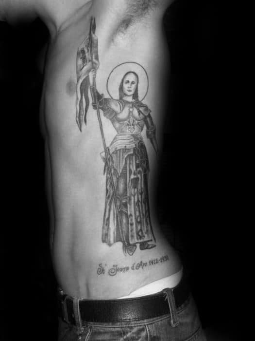 Black and grey Joan of Arc tattoo on the ribcage, featuring her in armor holding a flag with a halo around her head.