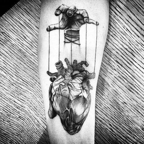 Realistic puppet tattoo of a human heart controlled by a hand with strings, symbolizing emotional manipulation or control.
