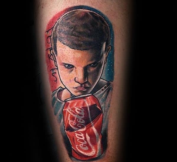Tattoo of Eleven from Stranger Things crushing a Coca-Cola can with her powers, intense gaze and vibrant red-blue background.