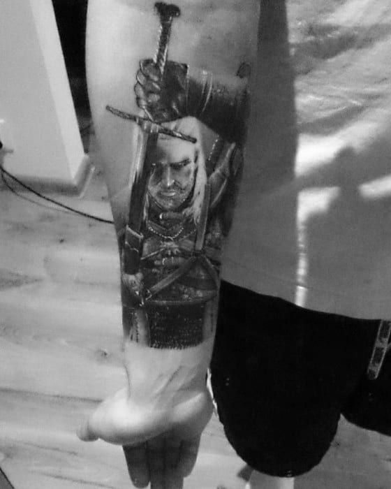 Realistic Witcher tattoo of Geralt holding two swords, positioned vertically on a man’s inner forearm in black and grey ink.