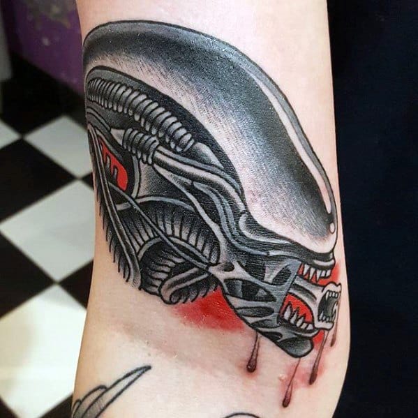 Stylized Xenomorph tattoo with bold black lines, red accents, and dripping blood effect on the forearm.