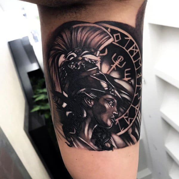 Dark-themed Athena tattoo with intense shading, Spartan helmet, and circular rune-like symbols behind her head on upper arm