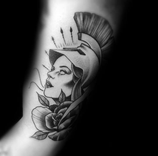 Stylized Athena tattoo with upward gaze, plumed helmet, rose detail, and fine black and grey shading on the upper arm