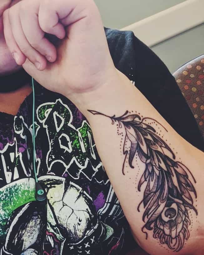 Black and grey peacock feather tattoo on the forearm, featuring intricate shading, fine details, and decorative dot accents.