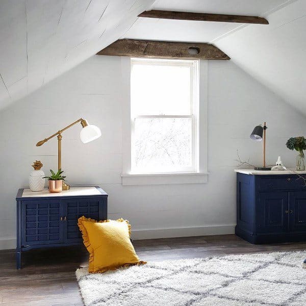 Cozy attic room with wooden beams, blue cabinets, a yellow pillow, and a window letting in natural light