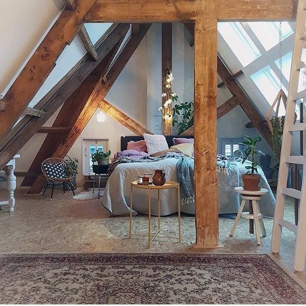Cozy attic bedroom with wooden beams, a large bed, lush plants, and soft lighting on a patterned rug