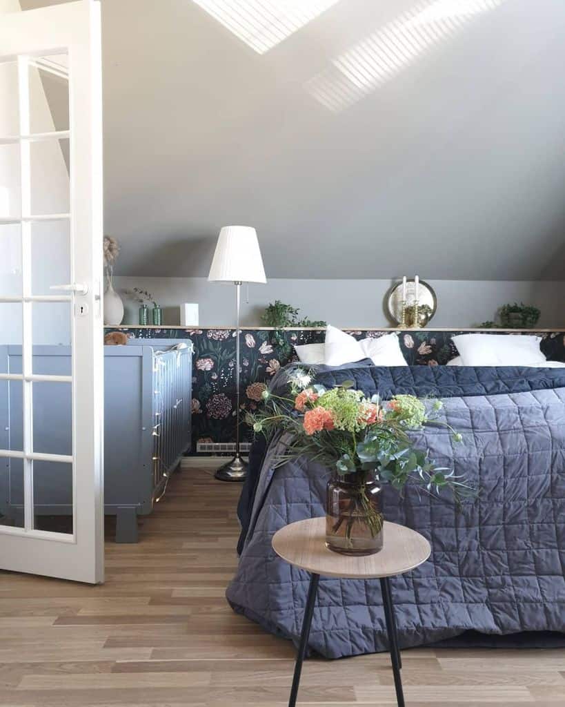 Cozy bedroom with floral wallpaper, a bed, table with flowers, and soft lighting from a tall lamp