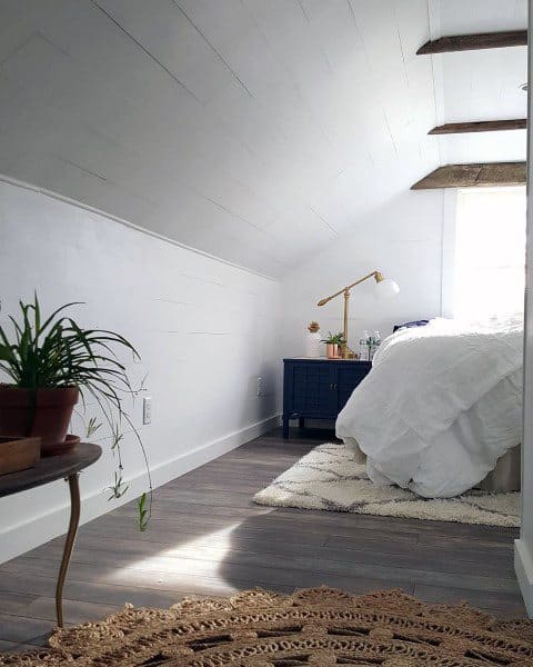 Cozy attic bedroom with a slanted ceiling, bed, potted plants, and a golden lamp on the navy nightstand by the window