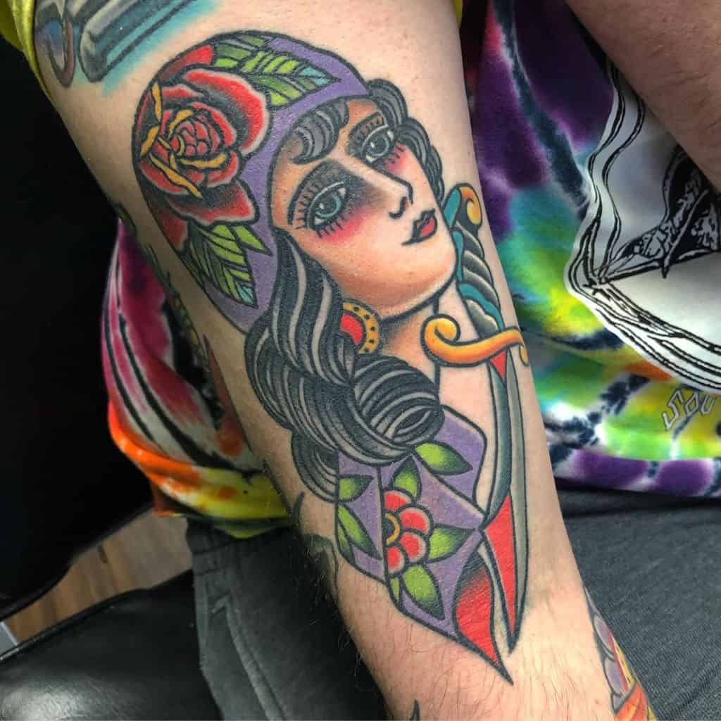Colorful traditional tattoo of a gypsy woman with a rose headscarf, bold shading, and intricate patterns on the forearm.