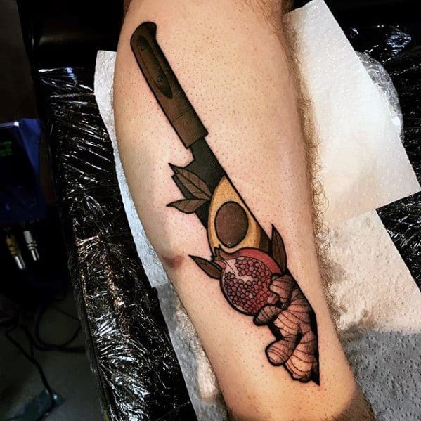 Bold avocado tattoo with a knife, pomegranate, and hand design, combining realism and illustrative style on the calf.