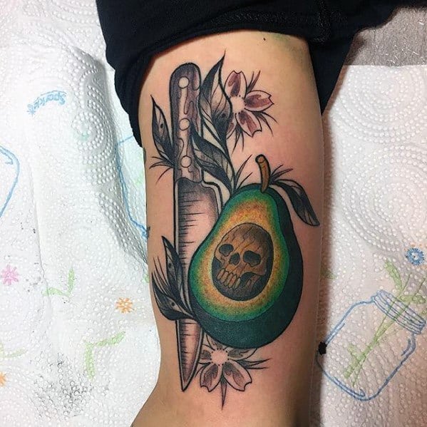 Colorful avocado tattoo with a skull seed, knife, and flowers, blending dark and botanical elements in a bold arm design.