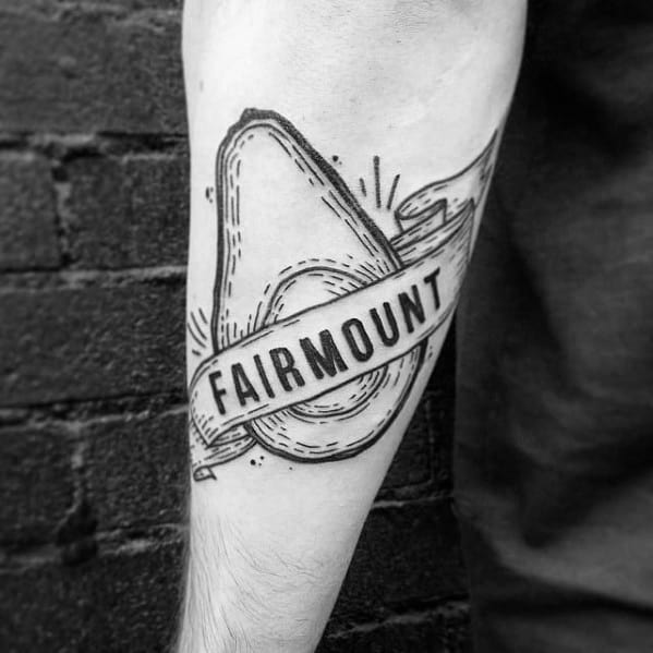Black linework avocado tattoo on the forearm with a banner reading “FAIRMOUNT,” styled with vintage-inspired shading and details.