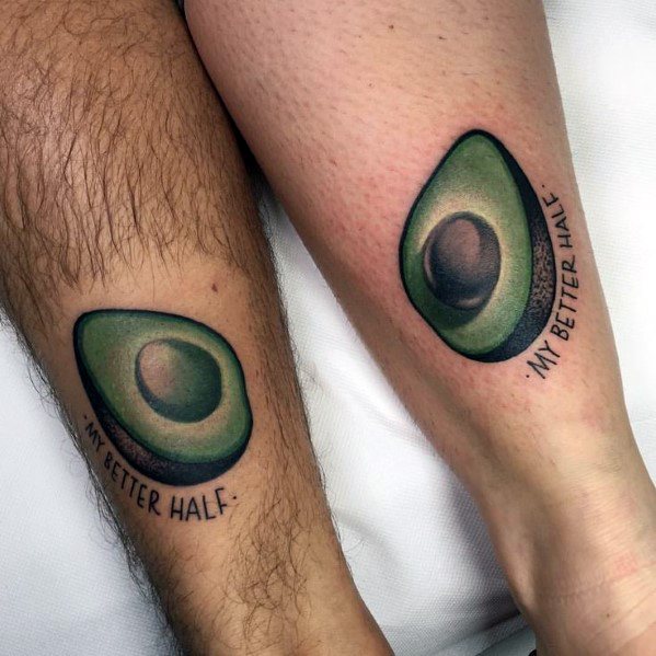 Matching avocado tattoos on two legs, each with a half and the phrase “MY BETTER HALF,” symbolizing love and connection.