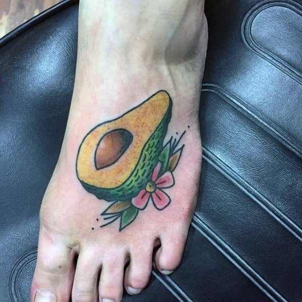 Colorful avocado tattoo on the foot with a pink flower and green leaves, featuring bold outlines and playful shading.