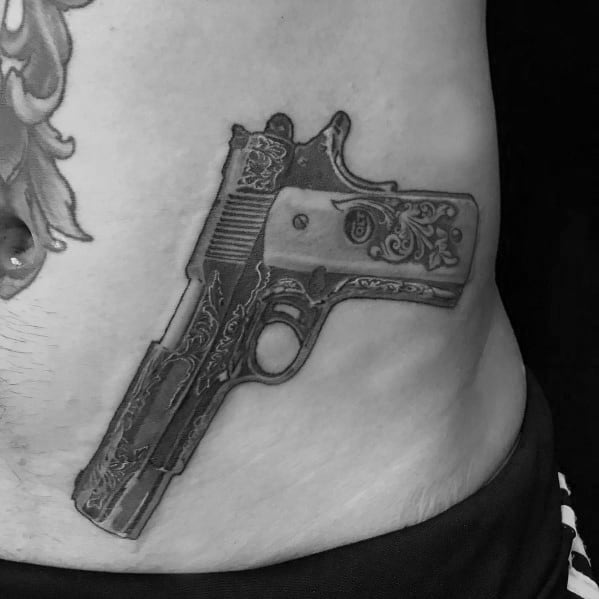 Realistic black and gray tattoo of an engraved 1911 pistol on the side of the torso, featuring intricate detailing for a bold and artistic look.