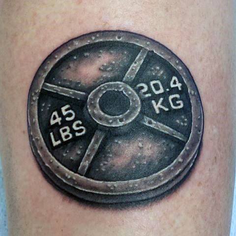 Realistic 45 lbs weight plate tattoo in black and grey, symbolizing dedication to strength training and personal growth.