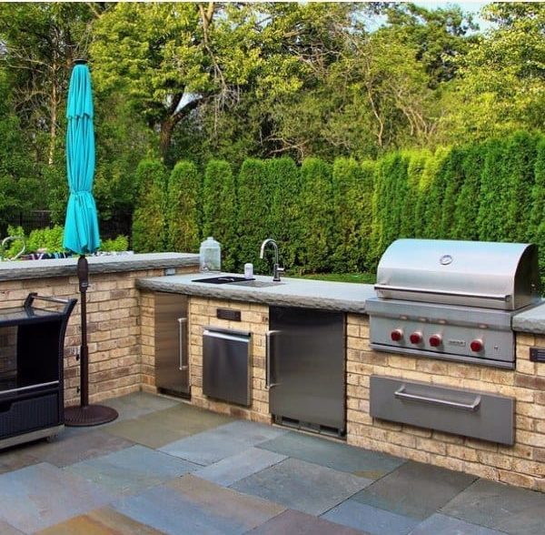 Outdoor kitchen with brick countertops stainless steel appliances and lush green hedge backdrop.