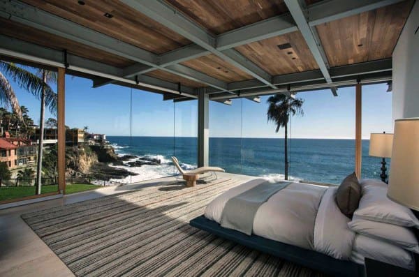 Beachfront bedroom with glass walls, ocean view, and wooden ceiling design.