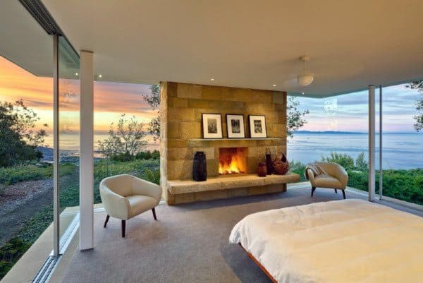Bedroom with glass walls, sunset ocean view, and a cozy stone fireplace.