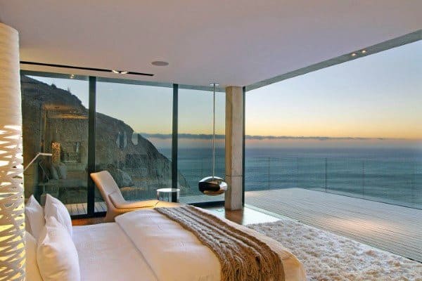 Modern bedroom with glass walls, ocean view at sunset, and cozy furnishings.