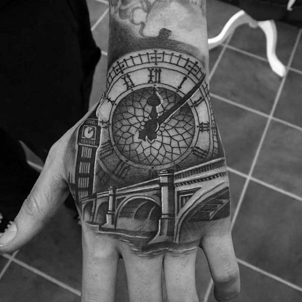 Detailed black and grey tattoo of Big Ben clock face on the hand, with intricate shading and the clock tower visible beneath.
