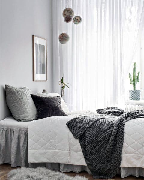 Minimalist bedroom with a white bed, grey pillows, knit blanket, potted cactus, and sheer curtains