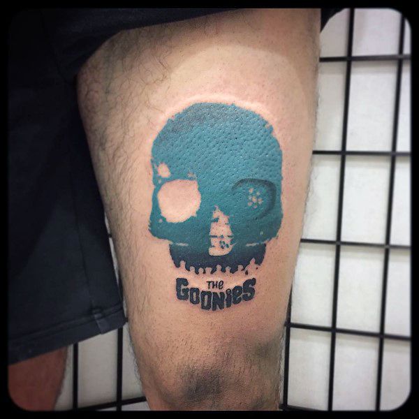 Minimalist tattoo of a blue skull from The Goonies, featuring a silhouette of a ship inside and the movie title in bold black text below.