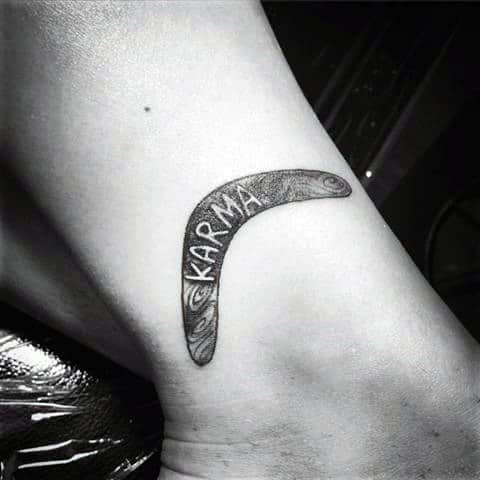 Minimalist black boomerang tattoo with the word KARMA and subtle swirling patterns on the inner arm.