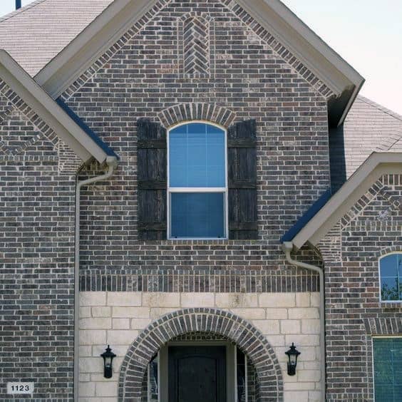 Charming house exterior featuring a mix of dark brick and light stone accents with arched entry and rustic wooden shutters