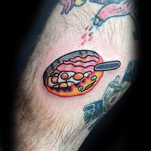 Calcifer tattoo with a frying pan on top, cooking bacon and eggs, featuring his classic wide-eyed, fiery expression.