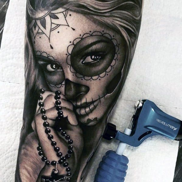 La Catrina tattoo of a woman with sugar skull makeup, holding a rosary with a cross, featuring intense eyes and fine shading.
