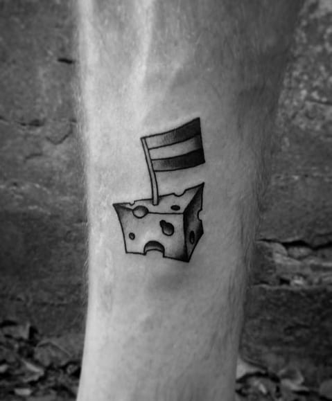 Small cheese block tattoo on the leg with a flag planted on top, featuring bold outlines and shading for a simple yet playful design.