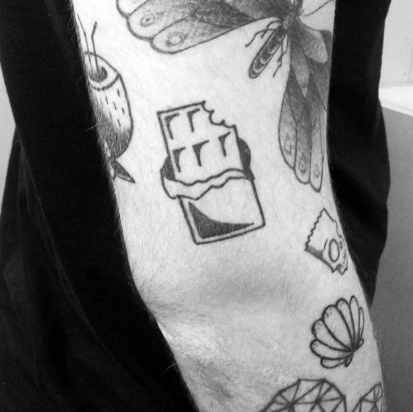 Minimalist black ink tattoo of a bitten chocolate bar in a wrapper, placed on the forearm among other small line-art tattoos.