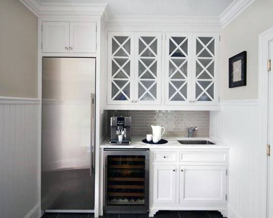 White coffee bar with glass cabinet doors, built-in fridge, coffee machine, and small sink.