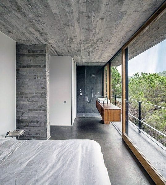 Modern bedroom with concrete floors, large windows, and a minimalist bathroom with a wooden vanity overlooking nature