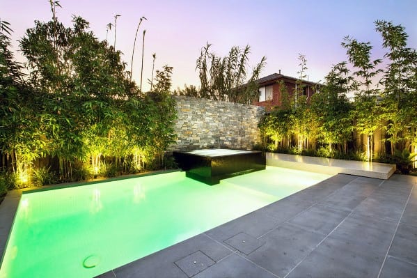 Modern backyard pool with illuminated green water, bamboo plants, and a stone waterfall feature at sunset