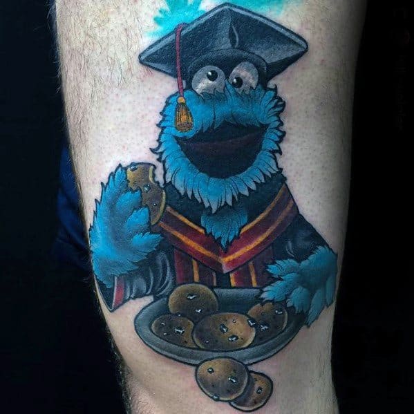 Graduation-themed Cookie Monster tattoo with a cap and gown, joyfully holding a plate full of cookies and one in his paw.