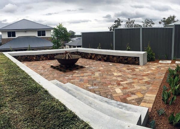 Brick patio with concrete seating, a central metal fire pit, and stacked wood storage.