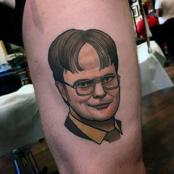 Realistic tattoo of Dwight Schrute with his signature glasses, brown hair, and mustard shirt, featuring smooth shading and bold outlines on a leg.