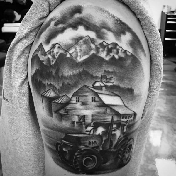 Black and gray upper arm tattoo of a tractor near a barn with silos, set against mountains and cloudy skies in the background.