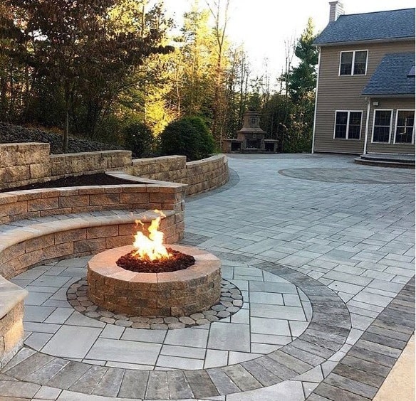A spacious backyard with a circular fire pit on a stone patio, encircled by trees and a two-story house