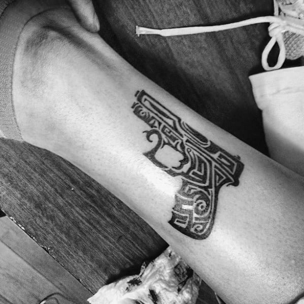 Black ink tattoo of a pistol with intricate tribal-style patterns, placed on the lower leg, combining firearm imagery with artistic design.