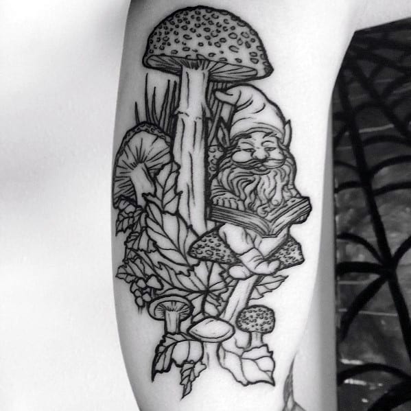 Black and white gnome tattoo featuring a bearded gnome reading a book while sitting on a mushroom, surrounded by large fungi and foliage.