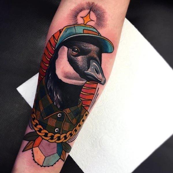 Colorful tattoo of a goose wearing a plaid shirt, gold chain, and teal cap, framed by geometric shapes, inked on a person’s forearm.