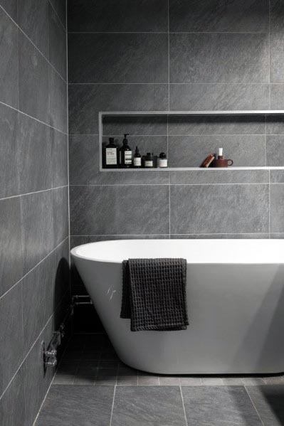 A modern bathroom with sleek gray tiled walls and floors, featuring a minimalist white freestanding bathtub and a built-in niche for convenient storage.