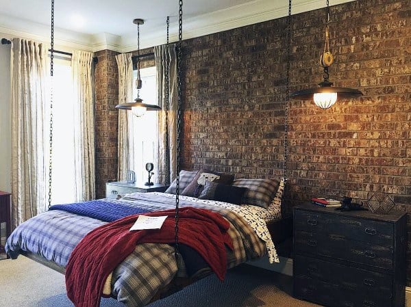 Industrial-style hanging bed with chains, plaid bedding, and exposed brick walls in a cozy bedroom.