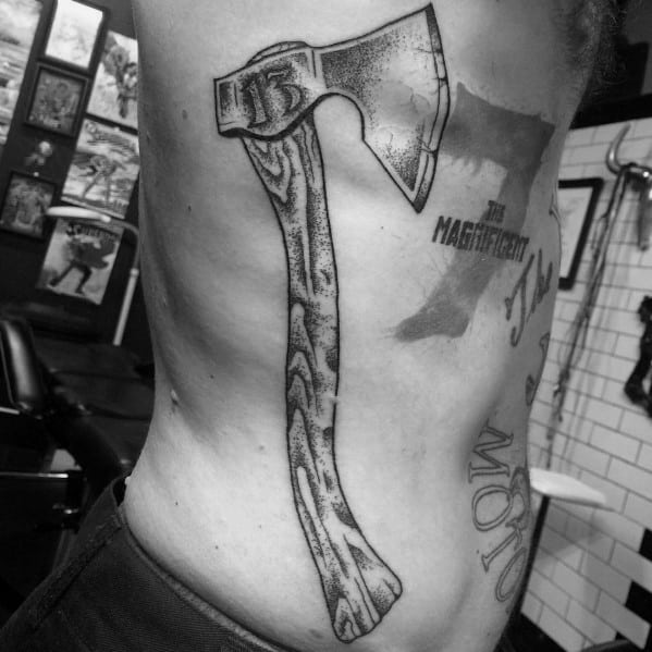 Detailed hatchet tattoo on the ribcage with wood grain texture on the handle and a rugged, shaded blade featuring the number 13.