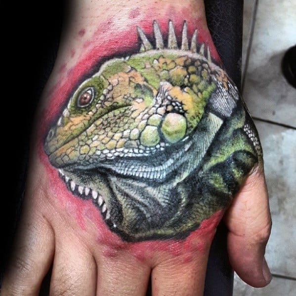 Colorful iguana head tattoo on hand with vivid green and yellow scales, sharp spines, and red background shading for contrast.
