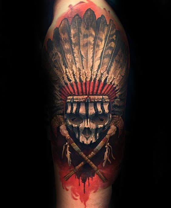 Tattoo of a skull wearing a feather headdress with tomahawks below it on an arm