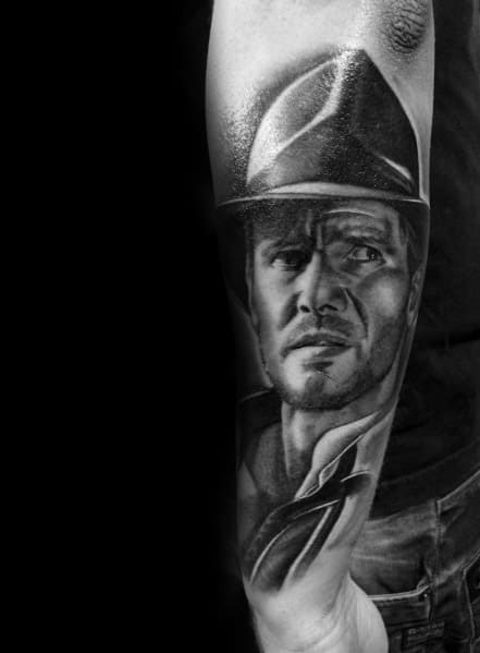Realistic black and gray Indiana Jones tattoo showcasing his intense gaze, iconic fedora, and rugged adventurer look.